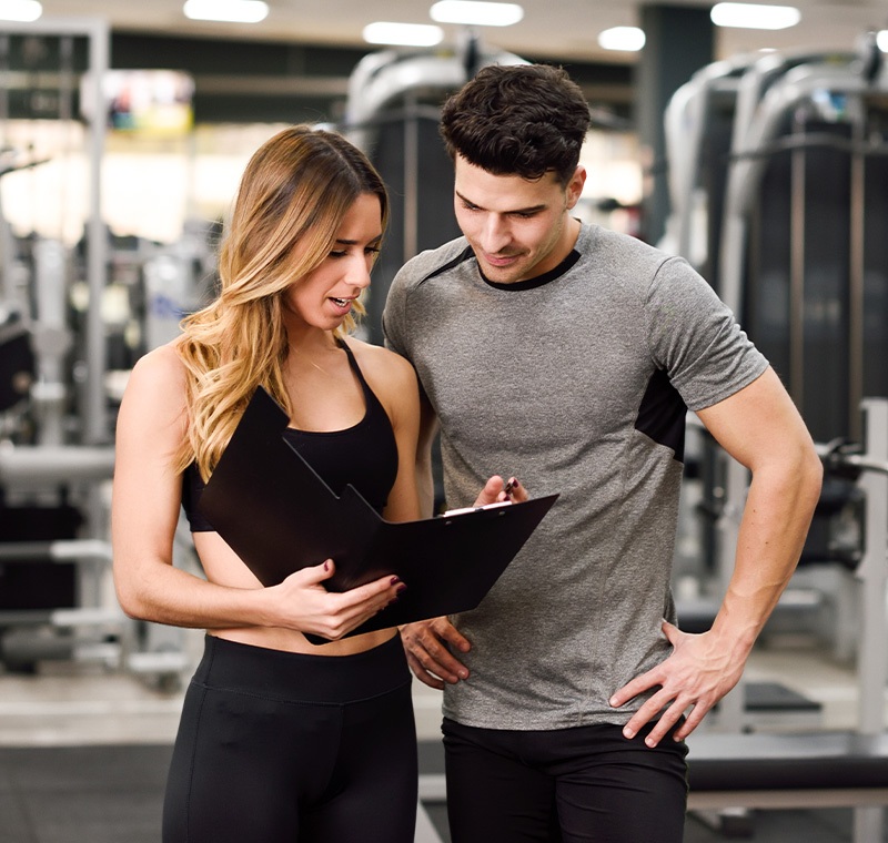 A smiling male fitness trainer in a gym environment shows a clipboard to a female client who holds a water bottle, both dressed in athletic wear and standing among weight benches and equipment.